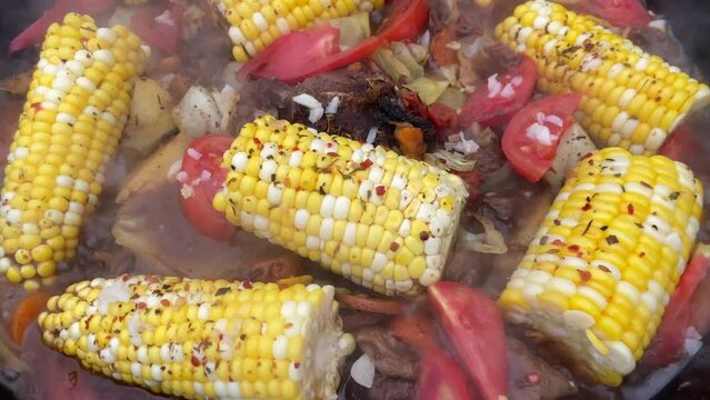 Corn with beef and vegetables. Shurpa in a cauldron. Tomato, red pepper and lamb soup cooked on hot coals. Hot beef hunting goulash or bograch soup with corn cobs, paprika, vegetables and spices.