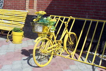 A yellow decorative bicycle with flowers stands in a parking lot on a summer day.