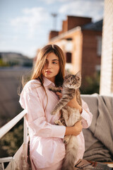 A girl with long hair in pink pajamas plays with a cat in the morning on the balcony.