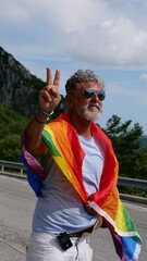 Portrait of a gray-haired senior elderly Caucasian man bisexuality with a beard and sunglasses with a rainbow LGBTQIA peace flag in mountains. Celebrates Pride Month, Rainbow Flag Day, gay parade