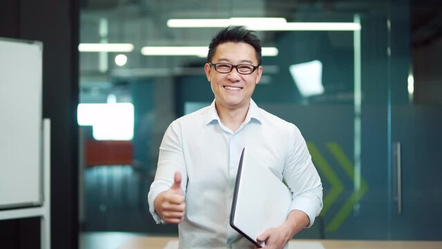portrait of young asian man specialist, employee programmer or teacher standing in modern office or classroom. Happy male wearing glasses is looking at camera and smiling indoors. thumb up gesture