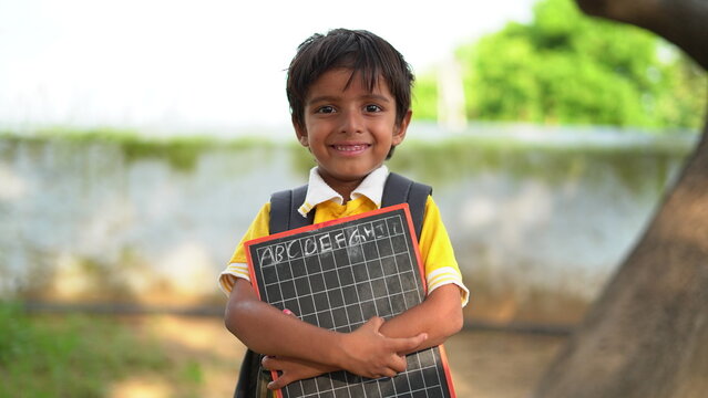 Portrait of happy cute little indian boy in school uniform holding blank slate against orange background, Adorable elementary kid showing black board. child education concept. rural india.