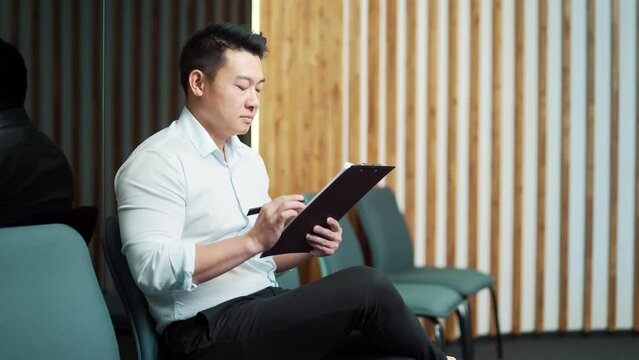 young asian male in waiting room for job interview or meeting. Thoughtful employee man holding folder paper while sitting at chair fills out a resume questionnaire in office. 