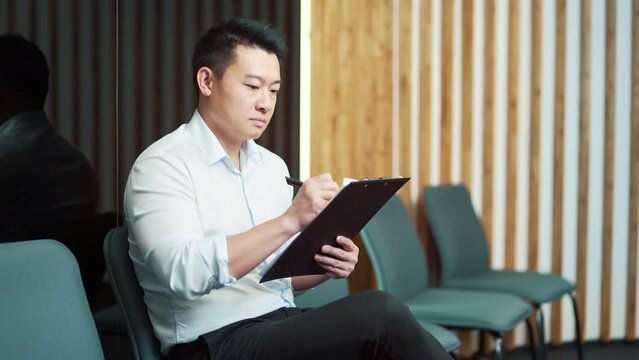 young asian male in waiting room for job interview or meeting. Thoughtful employee man holding folder paper while sitting at chair fills out a resume questionnaire in office. 