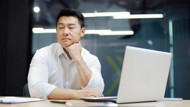 Bored asian office worker sitting at work workplace at computer desk leaning on hand. Tired man. Exhausted Businessman employee. Portrait Depressed, unhappy, workless male manager. indoor
