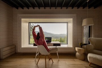 Woman stretching while works on laptop by the table in front of panoramic window with great view on mountains. Remote work and escaping to nature concept