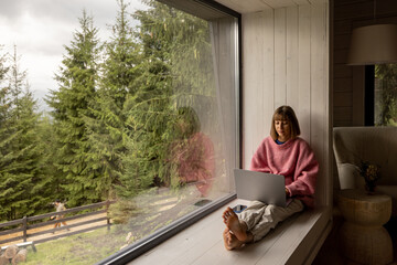 Woman works on laptop while sitting on a window with great view on mountains. Remote work from cozy...