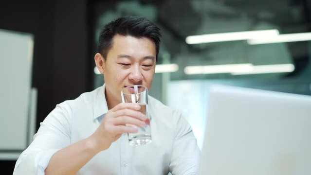 Asian employee man works in the modern office at the workplace sitting at the laptop computer drinking clean water from a glass. Business man, worker, quenches thirst Indoors