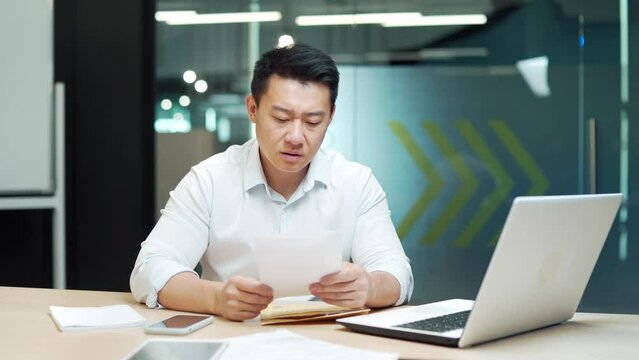 Worried Asian entrepreneur reading a letter with bad news Shocked frustrated business man received a rejection. male manager employee unpacking banking notification, law order document at workplace