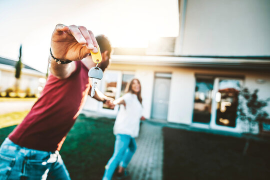 Happy young couple holding home keys after buying real estate - Husband and wife standing outside in front of their new house