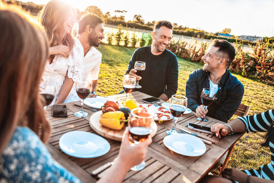 Group of friends having fun at bbq outside dinner in home garden - Happy people cheering red wine sitting outdoor at dining table - Social gathering, youth, food and beverage concept