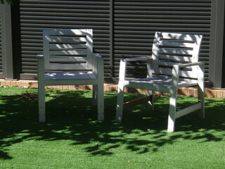 dos sillas blancas, situadas entre sol y sombras, en jardín interior, sobre césped artificial...