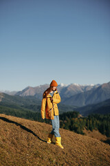 Woman autumn full-length walk on the hill smile with teeth and look at the mountains in a yellow raincoat and jeans happy hiking trip, freedom lifestyle 