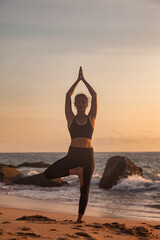 Fototapeta na wymiar Silhouette young woman on tropical sea coast sandy beach does asana yoga position arms raised outdoors at sunset. Female performs exercises for healthy lifestyle to restore strength, spirit