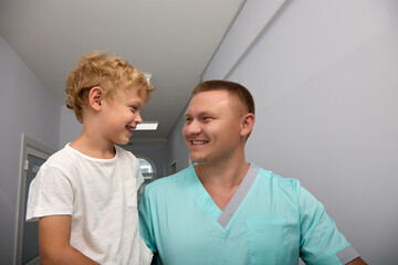 Smiling doctor holds the boy in his arms, brings him into the treatment room of the children's polyclinic. Kind doctor takes care of his little patients in the hospital