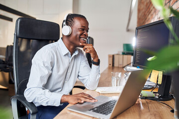 Laughing man programmer at office writing code while working with headphones in small office
