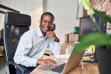 Portrait of smiling male programmer sitting by desk working on computer in small business office. Successful innovative African man working on laptop