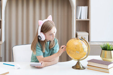 Fototapeta na wymiar Cute little girl in headset sitting at the desk and studying geography on the globe mockup. Home education concept