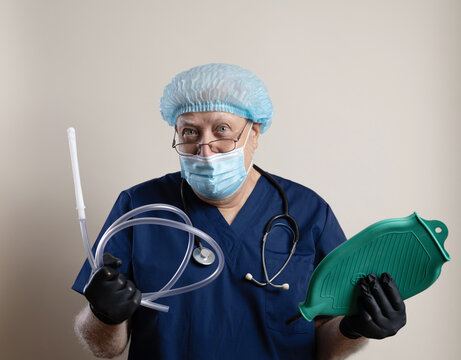 An elderly gray-haired doctor with a stethoscope and a cap holds an Esmarch Mug, looks into the camera, close-up, light background