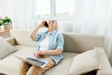 an elderly woman tired of work is sitting on a sofa in a cozy, bright apartment and talking on the phone with her head thrown up holding a laptop on her lap