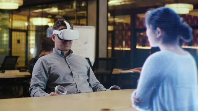 A man with virtual reality headset sits in an office table using joystics. Takes interview of a candidate using Augmented Reality 3D Model Concept. Hologram of girl appears infront of the man.