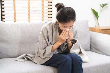 Fototapeta premium Sick, Coronavirus covid-19 asian young woman, girl headache under blanket have a fever, flu and use tissues paper sneezing nose, runny sitting on sofa bed at home. Health care on virus person.