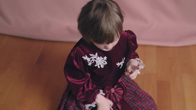 Cute little girl in an vintage burgundy dress is playing with a hair clip. The concept of children theater productions and developing acting circles. Pink background