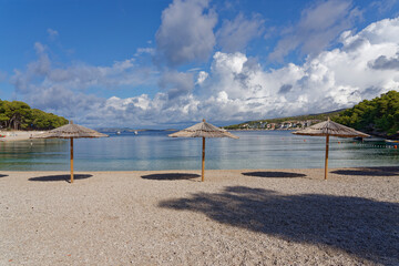 Beautiful beach on the Croatian Mediterranean coast with three reed sunshades in the foreground. In...