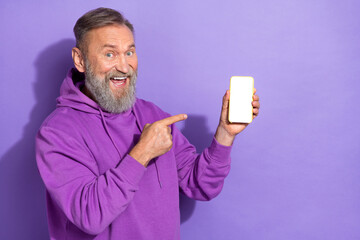 Photo of old handsome positive cheerful man wear purple hoodie directing at smartphone app ads isolated on violet color background