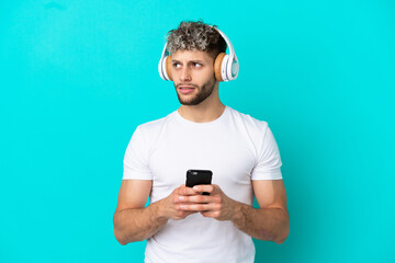 Young handsome caucasian man isolated on blue background listening music with a mobile and thinking
