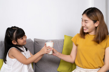 Young Asian mother hand over glass of fresh milk to her daughter inside of living room. Children who encourage to drink milk for good protein and nutrition.