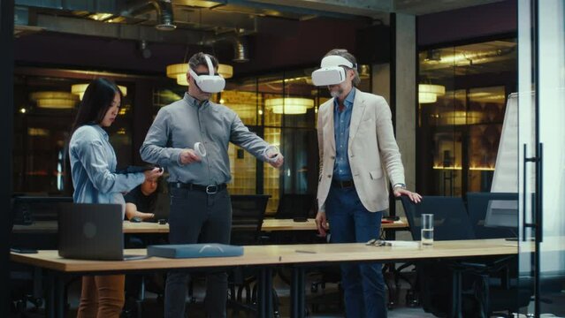 Multi-ethnic office workers testing VR headsets together, using tablet computer and wireless controllers, watching data, working in modern office of hi-tech company. Cyberspace digital technology