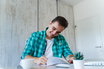 A young guy doing business work in his home office during the day while talking on phone using laptop and his mobile phone	
