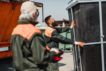 Young and senior garbage men working together on emptying dustbins for trash removal.