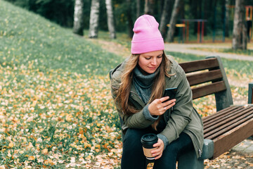 Woman sitting on outdoor in the forest drinking a cup of hot coffee and using a mobile phone technology. Beautiful female relax and enjoy outdoor activity lifestyle in nature on autumn	