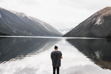 caucasian boy with long collected hair with his back to the lake and the mountains looking and...