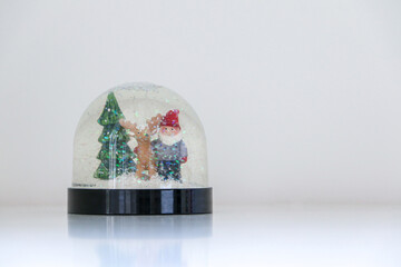 A sparkling and glittering Christmas snow globe with snow falling on Santa Claus, a reindeer, and a Christmas tree, on white snowy background, copy space on the right