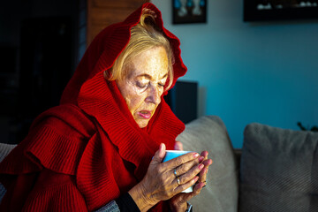 elderly lady drinking hot tea while freezing at home due to lack of heating; pensioners freezing at...