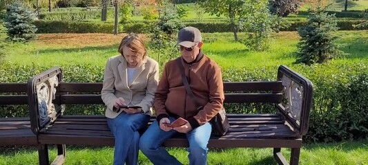 A couple of modern pensioners are sitting on a bench in the park with mobile phones and reading the news. An adult married couple with smartphones on a bench. The concept of modern pensioners.