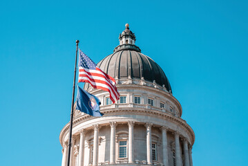 American flag waving at State Capitol Building in Salt Lake city. Low angle view of dome structure with blue sky in background. Famous national political landmark in city during summer.