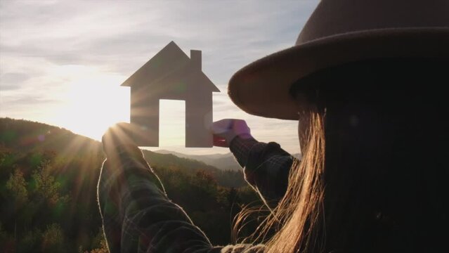 Close up of female hands holding small paper house against sunset or sunrise light on mountains background. Home in rays of sun in autumn nature. Family life, mortgage and business real estate concept