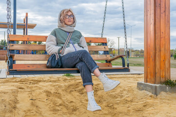 Young blonde woman in warm autumn clothes rides on wooden swing in recreation park. Evening walk in fresh air.