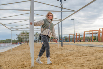 Young blond woman in warm autumn clothes is spinning around support of a metal structure in recreation park. Evening walk in fresh air.