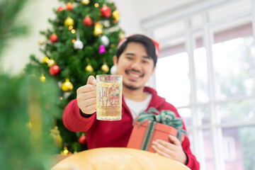 close up middle eastern man smile and raise glass of beer and hold gift box while sit on couch in living room with decorated of merry christmas tree for festival concept