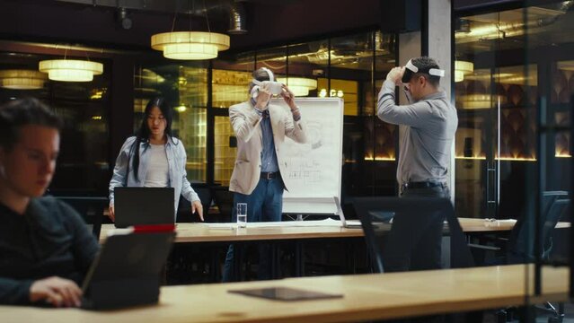 Multi-ethnic engineers in VR headsets, using laptop and wireless controllers, examining blueprint, watching data, working in modern office of hi-tech company. Cyberspace digital technology