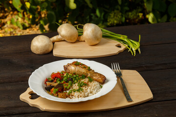 Rice with vegetables and chicken leg on a handmade wooden board next to champignons and green onions on a wooden table against a background of greenery.