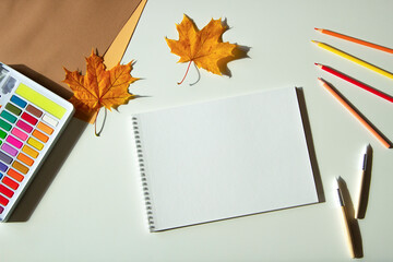 Autumn composition. Watercolor paint and brushes, open blank spirale sketchbook, colored pencils and autumn maple fall leaves on white desk background. Top view, copy space for text