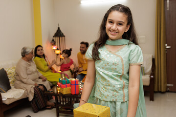 Fototapeta premium Young girl, kid, dressed up in ethnic wear holding gift boxes with smile expression pose for photo with family in background celebrating diwali Hindu festival Laxmi poojan