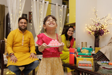 Obraz premium Cute little Kid, dressed up in ethnic wear holding plate of sweets with parents seating in the back expressing emotion looking at the candle celebrating diwali Hindu festival Laxmi poojan