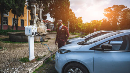 View of a fashionable bald bearded black man entrepreneur in a tailored dark-red suit just finished plugging his electric car into a street power supply; African guy next to an outdoor vehicle charger